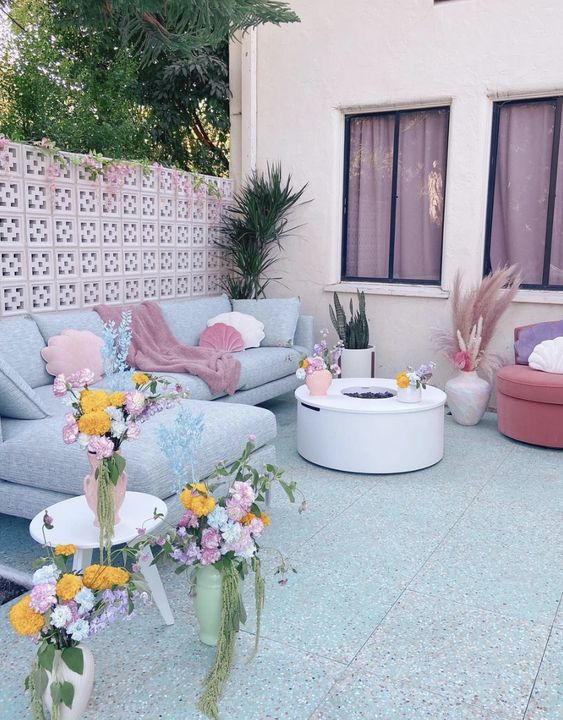 a dopamine decor terrace with a grey sofa and pastel pillows, a fire pit, potted plants and blooms and shell and cloud pillows