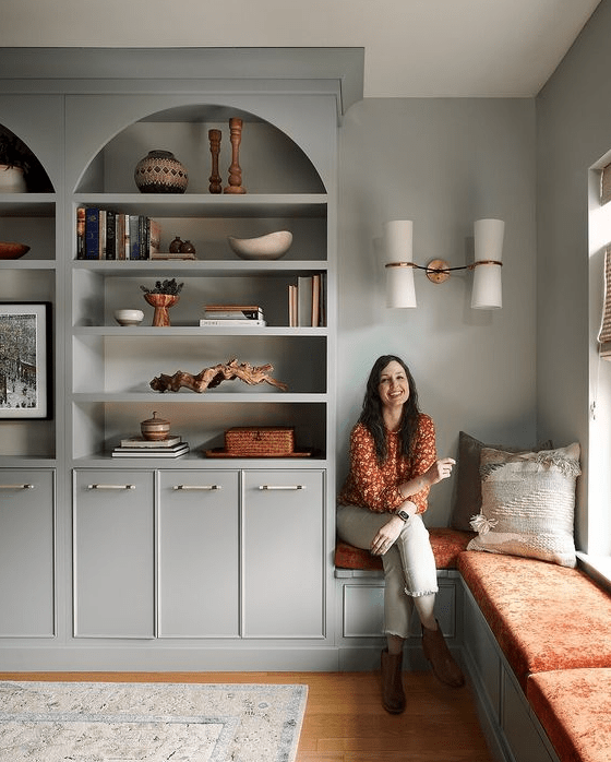 A dove grey living room with arched bookcases and built in cabinets, a windowsill bench, a wall lamp and bright upholstery