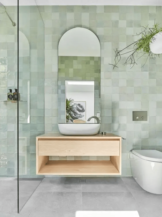 a dreamy and airy bathroom with green Zellige tiles on the walls, grey tiles on the floor, a floating vanity and an arched mirror