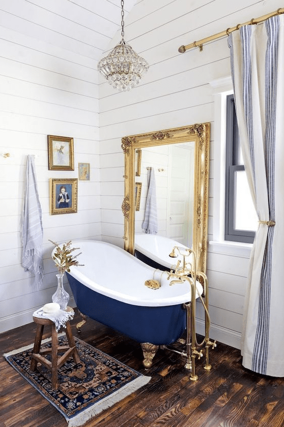 a fancy bathroom with white planked walls, a dark stained floor, a vintage navy bathtub, a floor mirror in a gilded frame, a mini gallery wall and a crystal pendant lamp