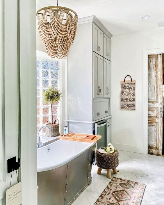 a farmhouse bathroom with a tub by the window, a wooden bead chandelier, a rug, a large storage unit and some boho decor