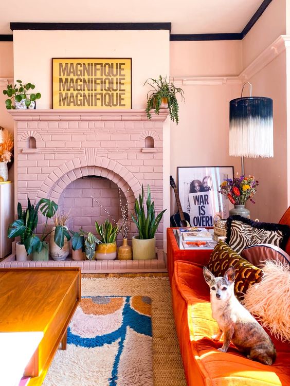 a fun dopamine decor living room with pink walls and a fireplace, an orange sofa with printed pillows, potted plants and a fringe lamp