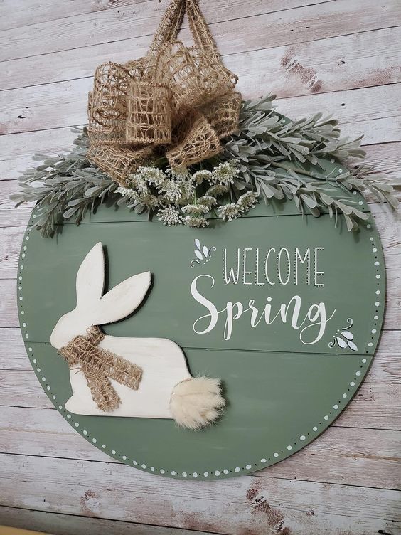 a fun spring sign in green, with a bunny wearing a scarf, faux greenery and blooms and a rustic mesh bow on top