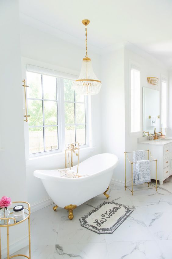 A glam white bathroom with windows, a free standing tub, a white vanity, a mirror and a chic and glam chandelier