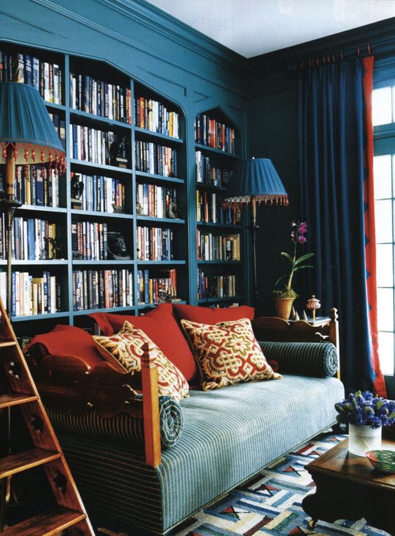 a home library of navy arched bookcases, a blue sofa with red pillows, blue floor lamps and a bold printed rug for a bold living room