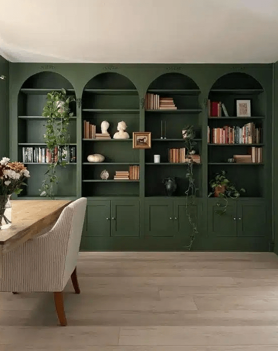 A home office with dark green built in arched bookcases, greenery, a desk and a white chair is a chic space