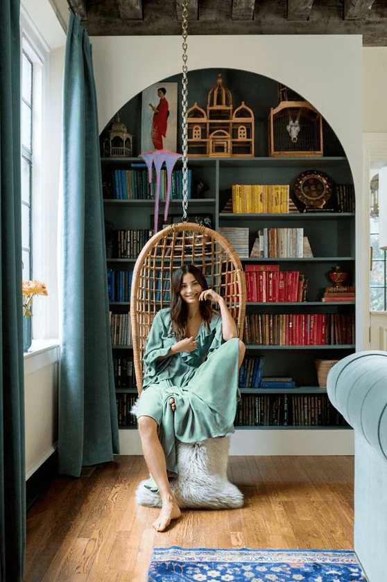 a large arched bookcase with dark green backing and shelves, colorful books and decor, a pendant chair to form a reading nook