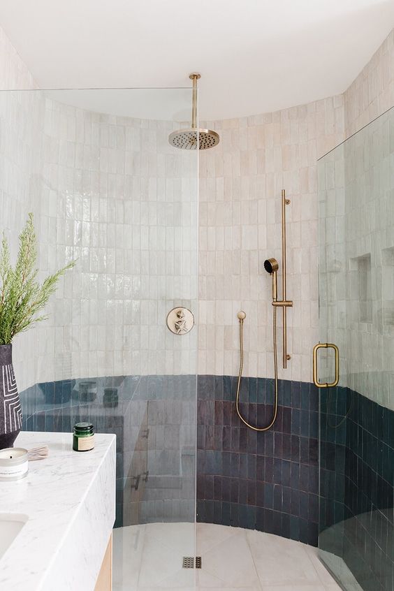 a large rounded shower space clad with skinny navy and white Zellige tile, a marble slab sink, brass hardware and greenery