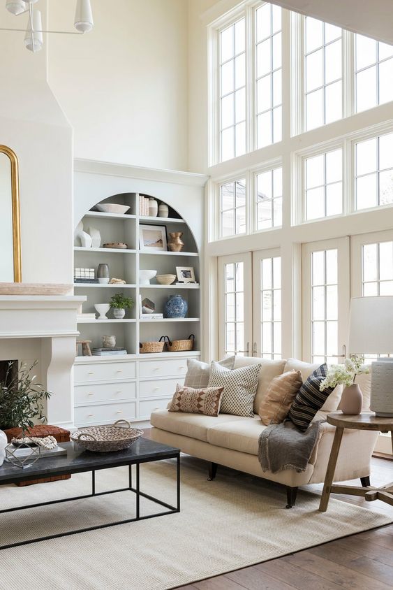 A light filled living room with an arched bookcase and a fireplace, a neutral sofa with pillows, a coffee table and some greenery