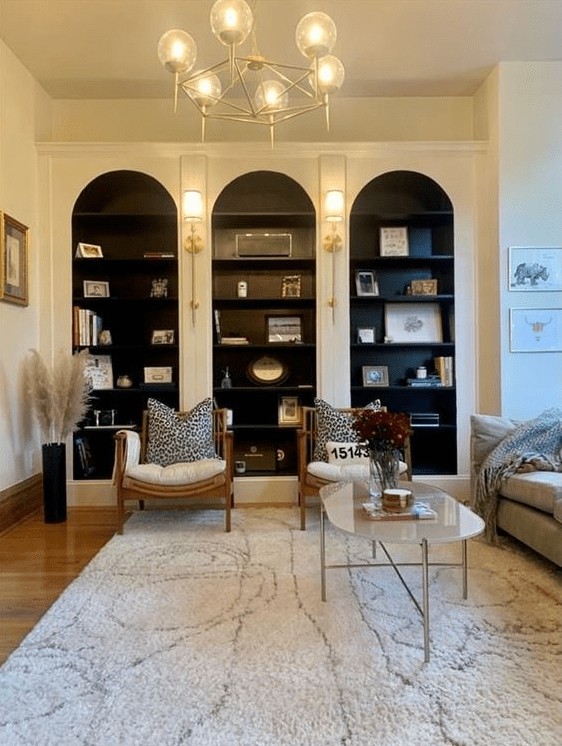 a living room with white arched bookcases with black shelves and backing, white chairs, a neutral sofa, a coffee table and a white rug