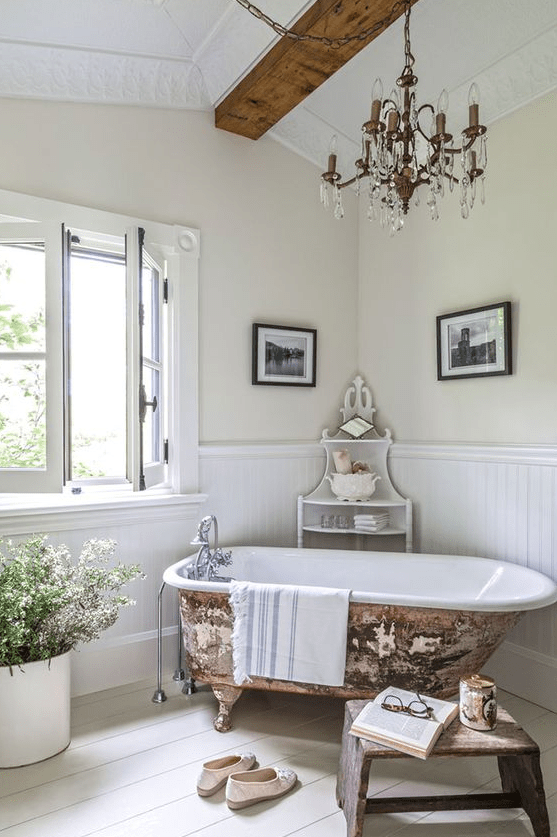 a lovely French country chic bathroom with wall paneling, wooden beams, a vintage chandelier, a clawfoot bathtub, a wooden bench and some blooms