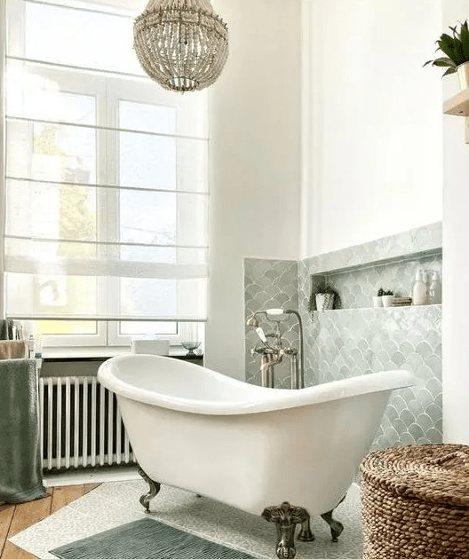 A lovely bathroom with a pale green fish scale backsplash, a free standing tub, a basket and some rugs