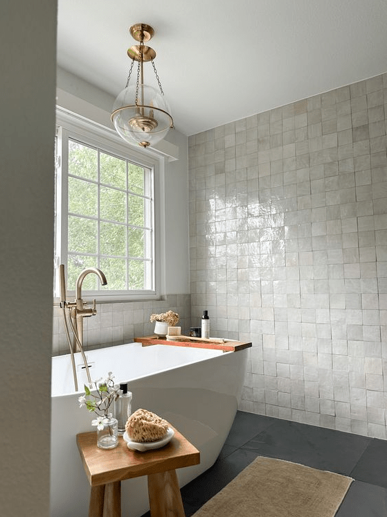 a lovely modern bathroom with a neutral Zellige tile floor, a grey floor, a tub, a wooden stool and a pendant lamp