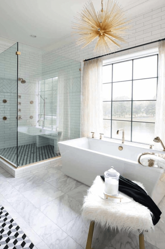 a lovely neutral bathroom with white and white marble tiles, a shower space, a tub by the window, a sunburst chandelier, a stool and brass fixtures