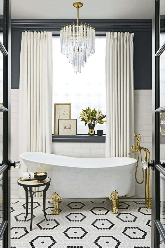 a luxorious bathroom with a black and white tile floor, white subway tiles and black walls, a chic tub, gold fixtures and a lovely chandelier