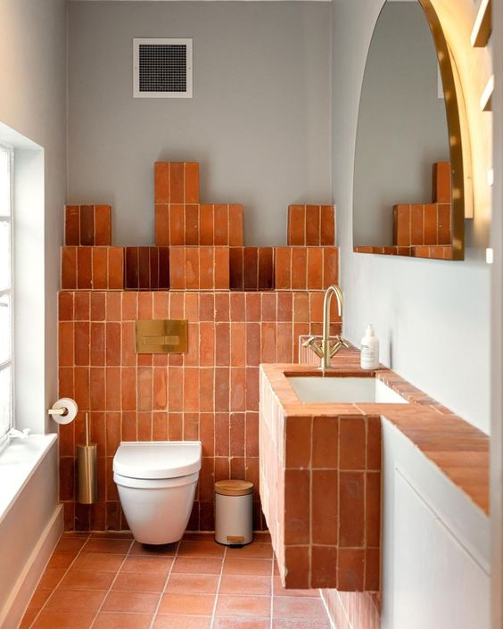 A minimal rustic bathroom clad with terracotta tiles, a built in sink, a semi circle mirror and a toilet