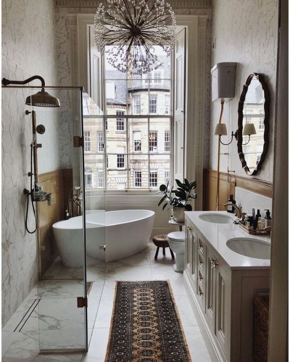 a modern Parisian bathroom with a window, a tub, a shower space, a white vanity, a mirror, a boho rug and a lovely flower-inspired chandelier