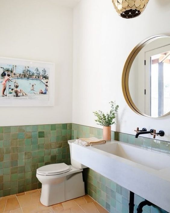 a modern bathroom done with green Zellige tile, a concrete wall-mounted sink, a toilet, a gold pendant lamp and a round mirror