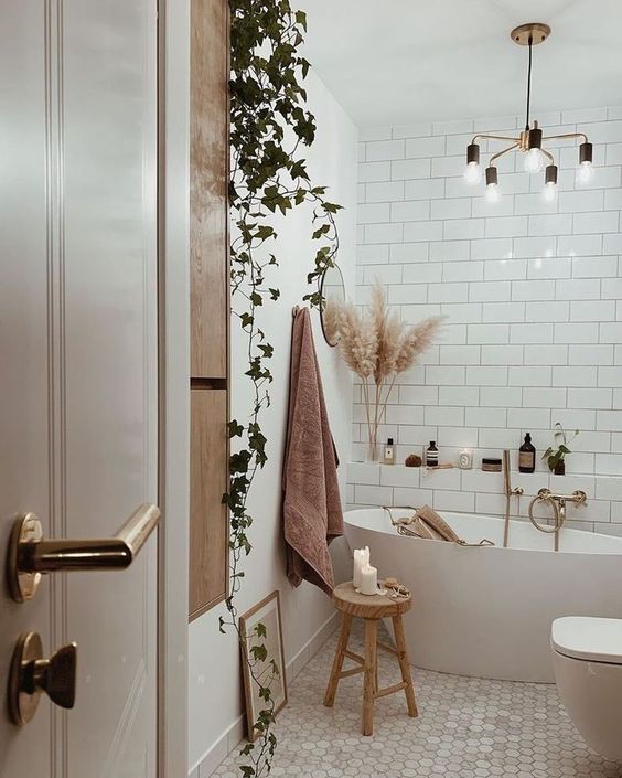 a modern boho bathroom done with white subway and hex tiles, a tub, a shelf with supplies, a modern chandelier and greenery
