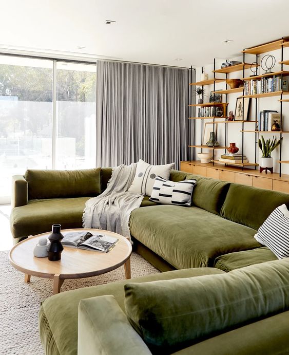 a modern living room with a storage unit with open shelves, a large green sectional, a coffee table, grey curtains and printed pillows