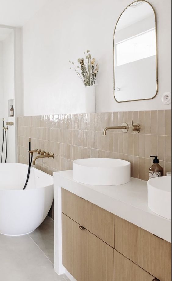 a modern rustic bathroom done with white walls, tan Zellige tile, a built-in vanity with drawers, a tub, round sinks and a mirror
