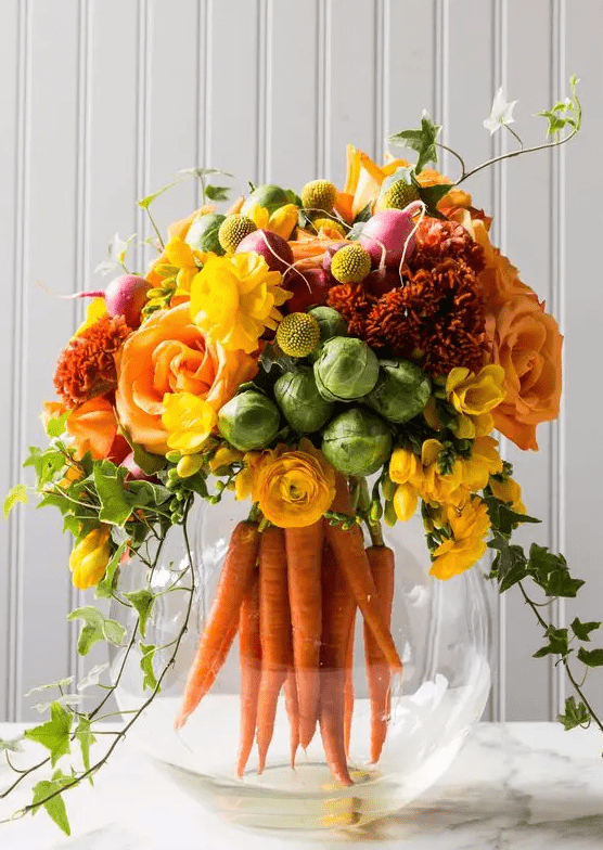 a modern rustic spring centerpiece of an aquarium with carrots, bold blooms and cabbages plus some foliage is a fun idea