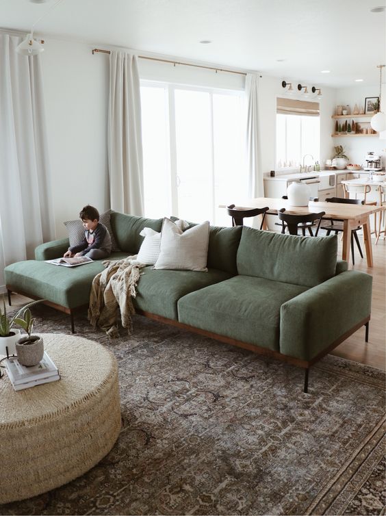 a modern space with a kitchen and dining zone and a living room with a green sofa, a coffee table, a rug and neutral curtains