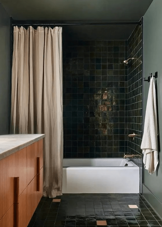 a moody bathroom with dark green walls and dark green square tiles, a timber vanity, some neutral textiles and gold and brass fixtures