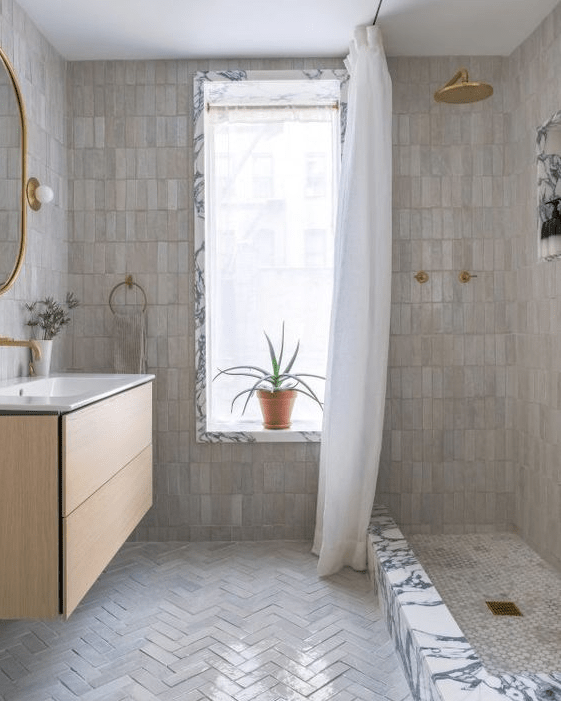 a neutral bathroom done with stacked Zellige tiles and herringbone ones, a shower, a floating vanity and gold fixtures