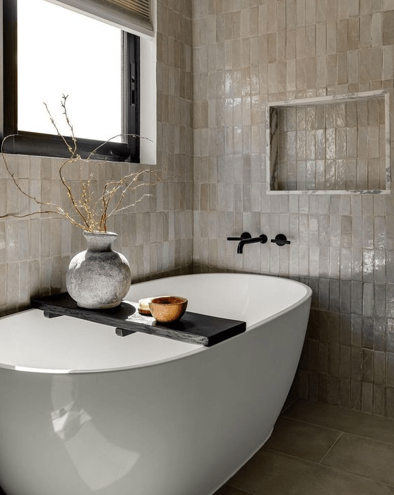 a neutral bathroom with stacked Zellige tiles, an oval tub, a niche, some branches in a vase and a window