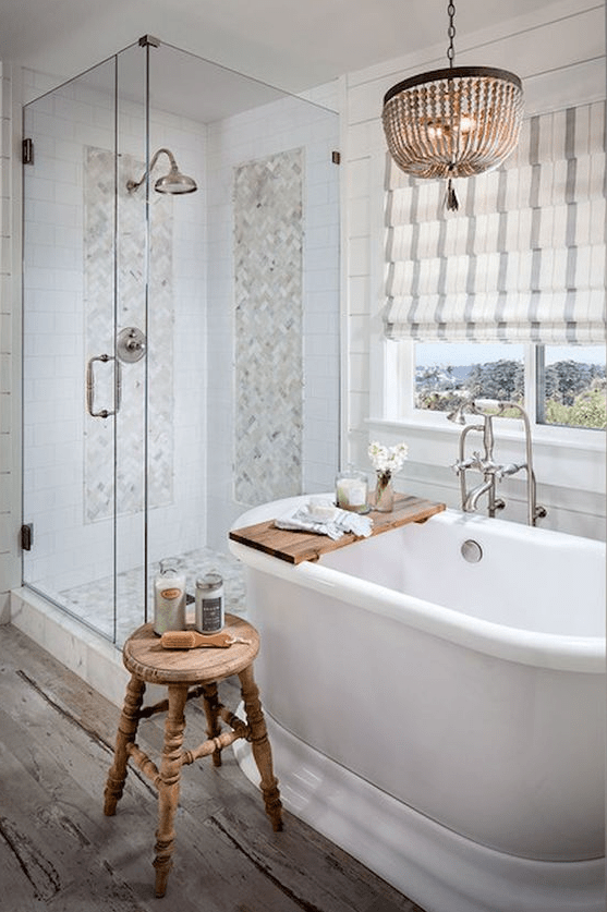 a neutral farmhouse bathroom with marble tiles, Roman shades, a bead chandelier and a vintage wooden stool
