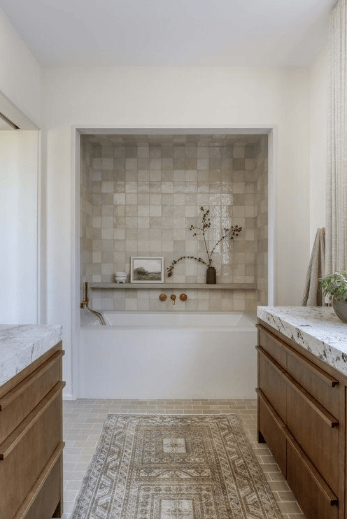 A neutral organic bathroom with a built in tub in a niche clad with Zellige tiles, rich stained vanities and white stone countertops