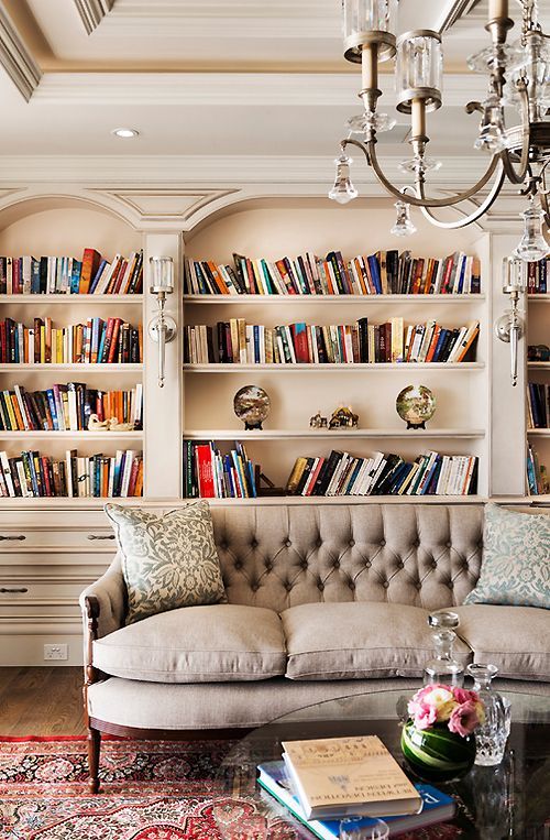 a neutral refined living room with arched niches with books, a grey sofa with pillows, a crystal chandelier and a printed rug