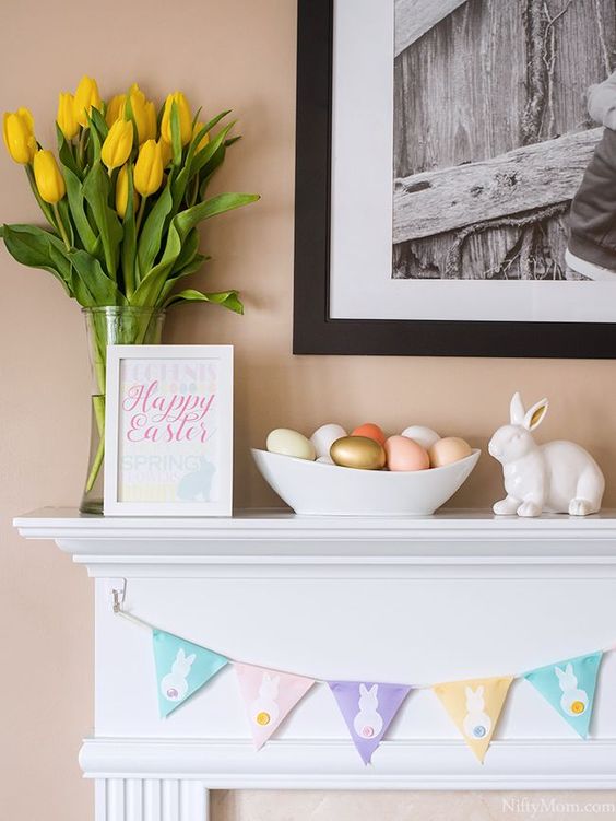 a pastel Easter mantel with a pastel bunny banner, some painted eggs, a bunny and yellow tulips is a cool and elegant idea