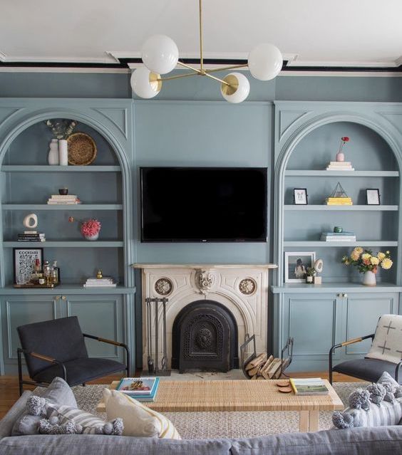 a pretty living room with pastel blue arched bookcases, a fireplace, a woven bench, mismatching chairs and a grey sofa