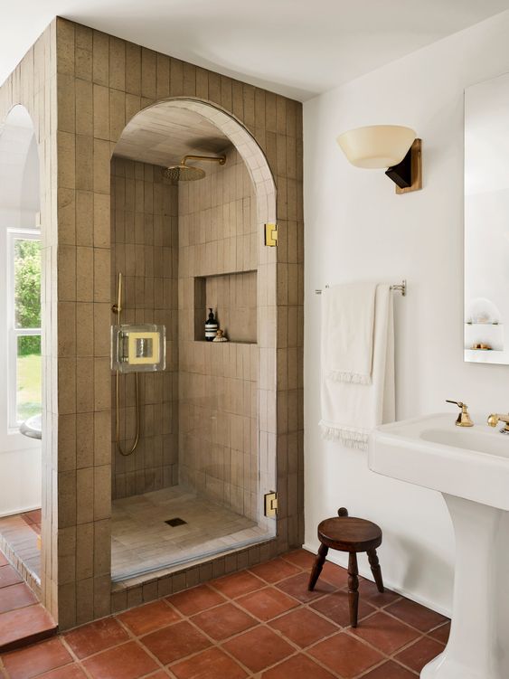 A refined bathroom clad with grey Zellige tiles and terracotta ones on the floor, a creative shower space, a free standing sink and gold touches