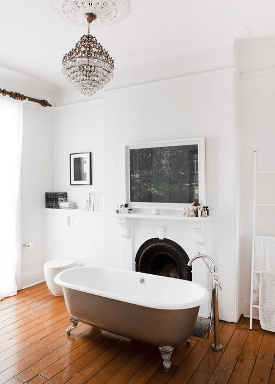 A refined bathroom with a rich stained wooden floor, a non working fireplace, lovely artworks, a crystal chandelier and a lovely bathtub