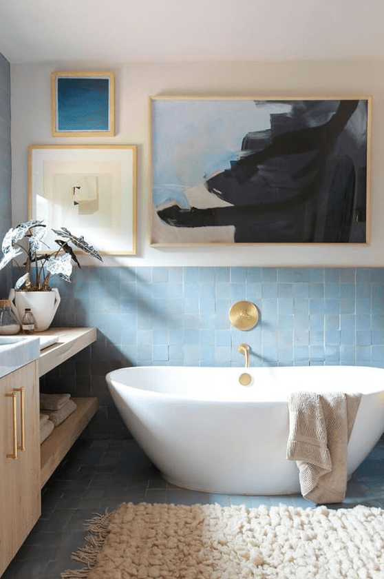 A refined bathroom with white walls, blue Zellige tiles, an oval tub, light stained furniture, a gallery wall and gold fixtures