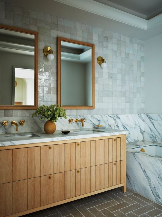 a refined neutral bathroom with white Zellige tile, a tub clad with marble, a stained vanity and brass hardware and fixtures