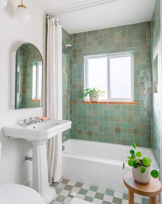 A retro bathroom with green and tan Zellige tile in the tub space, a window, a free standing tub, a stool with a potted plant and a shower curtain