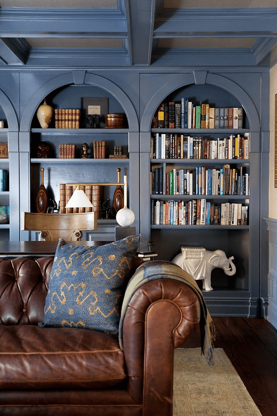 a row of navy arched bookcases, a desk and a chair, a brown leather sofa, lamps and cool decor