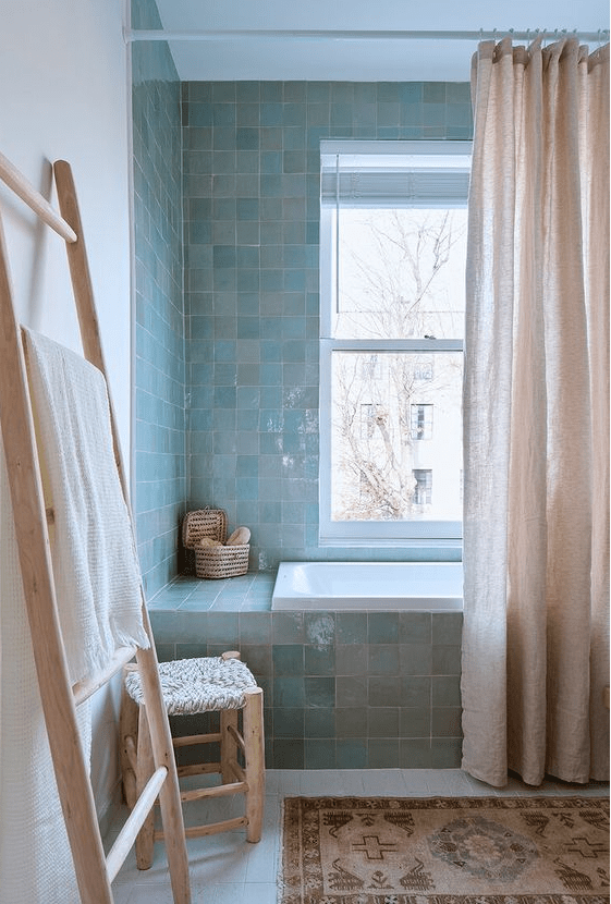 a serene bathroom clad with green and blue Zellige tiles, a tub with a pink curtain, a rug and some wooden furniture