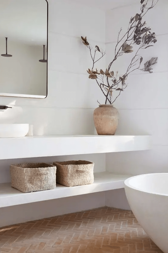 a serene white bathroom done with terracotta herringbone tile, an open vanity with baskets, a tub and some dried branches