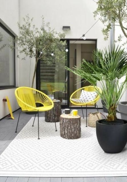 a small and bright patio with yellow chairs, stump side tables, potted plants and a printed rug is a great space