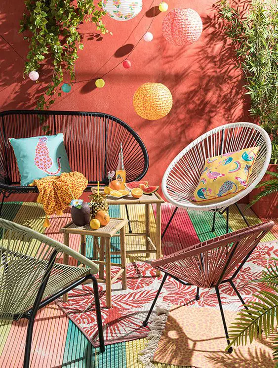 a small and colorful patio with a red wall, a loveseat and chairs, a duo of coffee tables, colorful rugs and lights