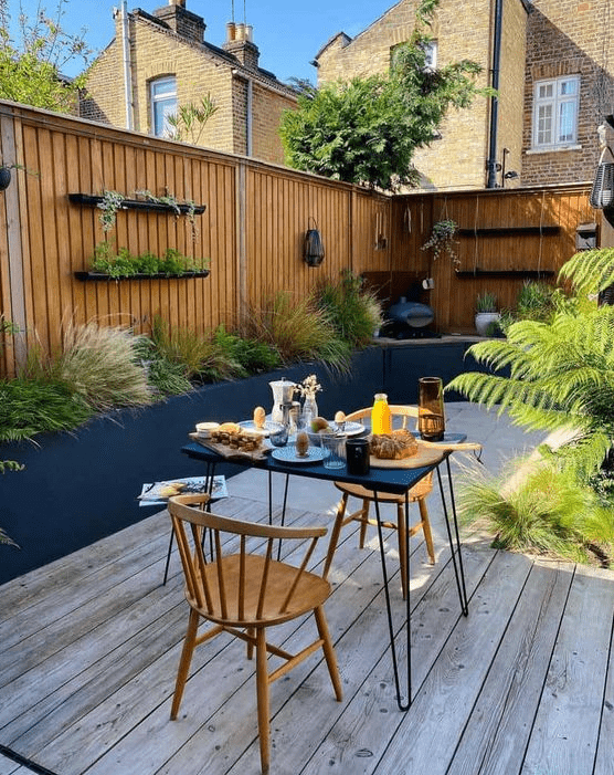 a small and cool patio with a wooden deck, greenery and grasses, a hairpin table and wooden chairs, some shelves is awesome