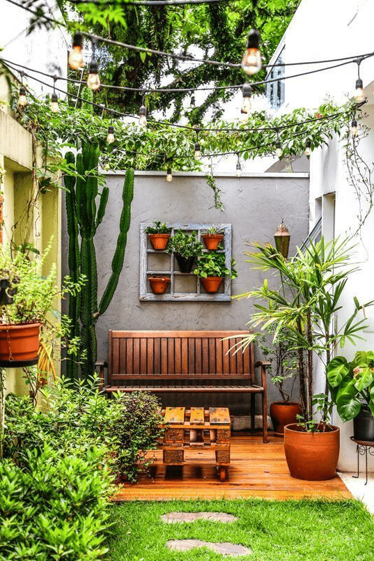 a small and cool patio with greenery, potted cacti and plants, a wooden bench and a pallet table, some lamps around