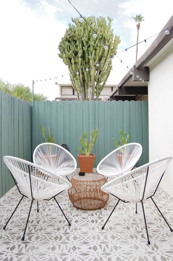 a small and cozy patio clad with tile, with white chairs, a coffee table and potted cacti plus string lights over the space