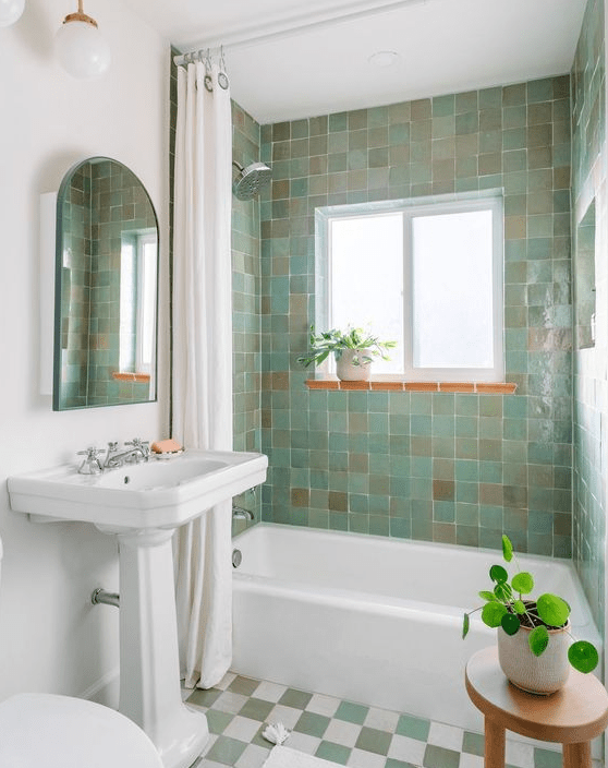 A small and cute bathroom with green and beige Zellige tiles, a checked floor, a free standing tub, some potted plants