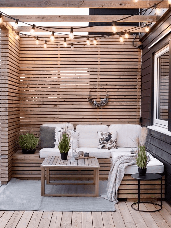 a small and cute patio with wood slat privacy screens and a bench, neutral cushions, a coffee table, string lights and potted greenery